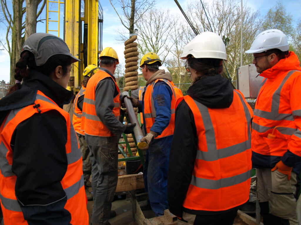 Five researchers and engineers in orange vests are looking at the drilling at the Delft demo site.